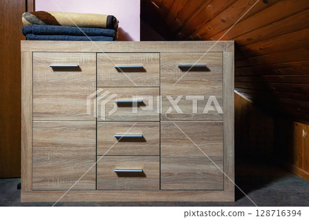 Wooden chest of drawers with folded blankets on top in small attic bedroom, front view Wooden chest of drawers with folded blankets on top in small attic bedroom, front view 128716394