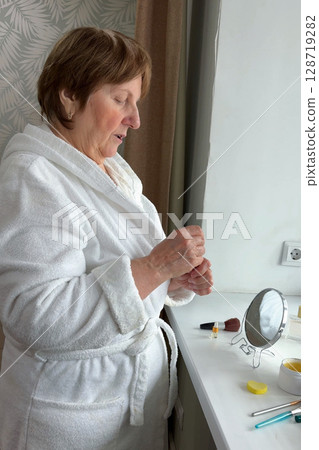 A beautiful senior woman is sitting at home and doing her make-up morning routine. Senior woman putting blush on her face 128719282