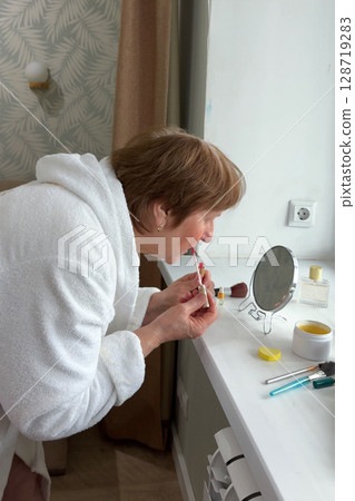 A beautiful senior woman is sitting at home and doing her make-up morning routine. Senior woman putting blush on her face 128719283