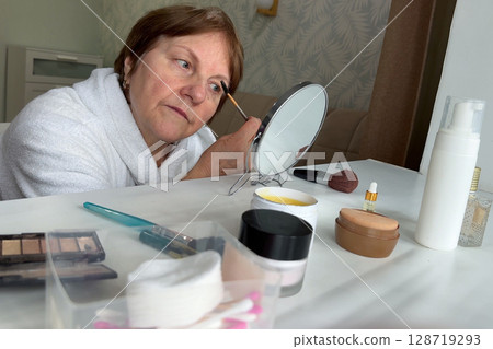 An older woman with charm is sitting in her living room, doing her makeup routine. 128719293