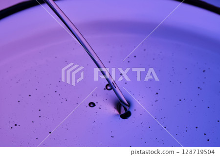 A jet of facial oil flowing into a petri dish on a purple background. A jet of facial oil flowing into a petri dish on a purple background. 128719504