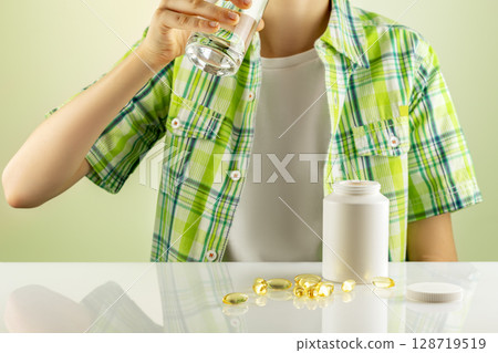 Organic kids vitamins and nutrients. Mockup of natural bio pills, vitamins or supplements. The boy washes down vitamins in gel capsules with water. Supplements of Omega 3 or D 3 Organic kids vitamins and nutrients. Mockup of natural bio pills, vitamins or supplements. The boy washes down vitamins in gel capsules with water. Supplements of Omega 3 or D 3 128719519