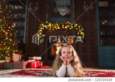 Merry Christmas. Little girl smiling near Christmas tree in classical dark interior. Young happy kid wearing deer horns in living room with fireplace Christmas tree gift boxes. Christmas eve at home Merry Christmas. Little girl smiling near Christmas tree in classical dark interior. Young happy kid wearing deer horns in living room with fireplace Christmas tree gift boxes. Christmas eve at home 128720326