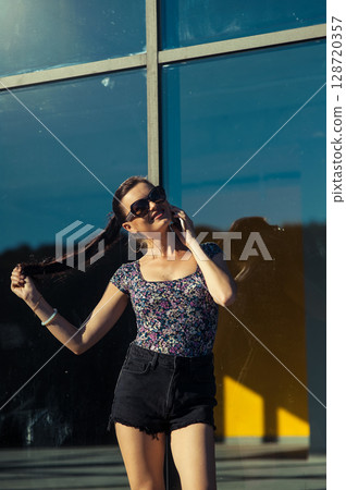 A girl wearing glasses is talking on the phone, leaning against the glass wall of a modern building A girl wearing glasses is talking on the phone, leaning against the glass wall of a modern building 128720357