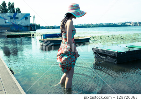 A young girl in a long colorful dress and a hat stands barefoot in the clear water of the lake A young girl in a long colorful dress and a hat stands barefoot in the clear water of the lake 128720365