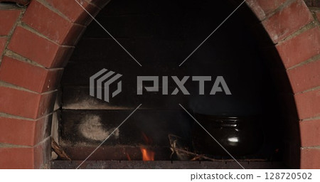 Clay pot placed inside rustic oven with vibrant flames growing steadily, showcasing traditional cooking techniques in an earthy environment that emanates warmth and comfort 128720502