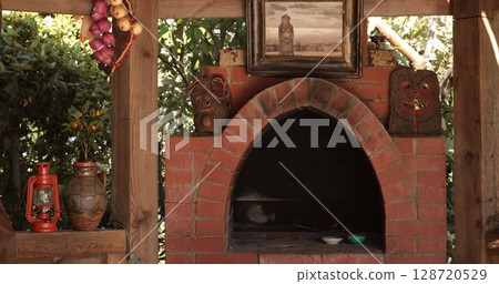 Rustic oven built with handmade bricks surrounded by natural elements including a vibrant green garden, vintage decor, artwork on the wall, and traditional pottery arranged nearby 128720529