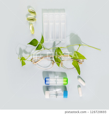 Ophthalmology concept with eyeglasses, containers with contact lenses, vitamins for eye health and moisturizing eye drops on blue background. Concept of eyecare, eye treatment and good vision. Flat la 128720693