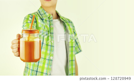 Boy, teenager holding carrot juice or vegetable smoothie in a glass mug with straw on a light background with copy space. Healthy food and drink for kid. Vegan or vegetarian diet 128720949