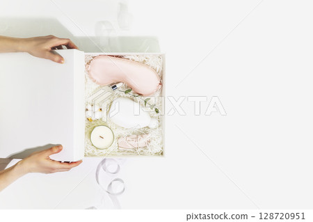 Female hands hold white gift box with sleep care set. Preparing self care package, craft gift box with mask for sleep, aroma candle and bubble bath. Relaxing at home and take care concept. Copy space 128720951