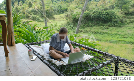 Young man typing on his laptop in a hammock, surrounded by lush greenery on a terrace of a bamboo hut, freelance and remote work in scenic natural setting Young man typing on his laptop in a hammock, surrounded by lush greenery on a terrace of a bamboo hut, freelance and remote work in scenic natural setting 128721088