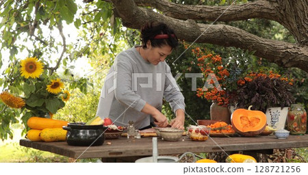 Woman prepares delicious Thanksgiving feast outdoors under leafy tree, surrounded by colorful autumn decor, fresh ingredients, and warm seasonal atmosphere 128721256