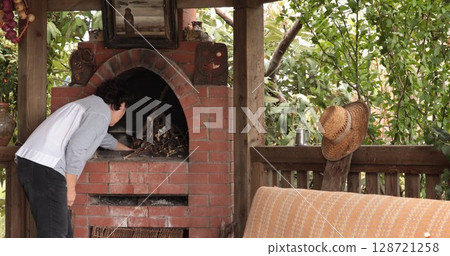 Outdoor oven set in garden shelter with person preparing wood for fire amidst greenery, showcasing rustic cooking atmosphere that encourages outdoor culinary experiences 128721258