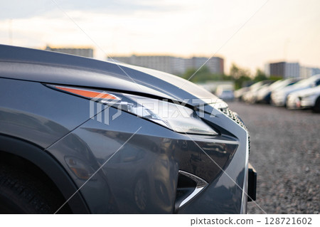 Close-up of the front of a gray car with the headlights on, parked in a lot with other cars in the background. 128721602