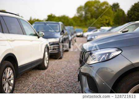 A row of various cars for sale outdoors, potentially at a car dealership. The vehicles are parked on a gravel surface with trees in the background. 128721613