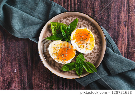 Close up of soft-boiled egg split in half with spices and basil leaves on a plate on a table top view Close up of soft-boiled egg split in half with spices and basil leaves on a plate on a table top view 128721658