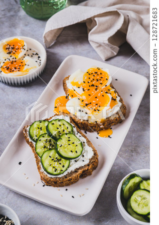 Cereal bread with cottage cheese, boiled egg and cucumber on a plate on the table vertical view 128721683