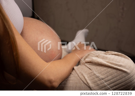 Pregnant woman sitting on a couch, touching, massaging painful ankle. Expectant mother tired of overweight, having health problems, muscles, joint pain. Cropped shot, close up Pregnant woman sitting on a couch, touching, massaging painful ankle. Expectant mother tired of overweight, having health problems, muscles, joint pain. Cropped shot, close up 128722839