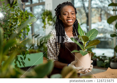 Cute african american woman in apron standing at the desk with plants Cute african american woman in apron standing at the desk with plants 128723875