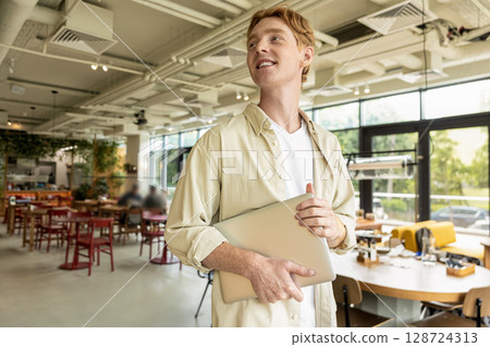 Positive young man with a laptop in hands standing in a cafe Positive young man with a laptop in hands standing in a cafe 128724313