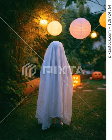 Child in ghost costume stands in glowing Halloween garden with lanterns and pumpkins. Ideal for spooky decor, festive blog posts, or autumn content Child in ghost costume stands in glowing Halloween garden with lanterns and pumpkins. Ideal for spooky decor, festive blog posts, or autumn content 128724564