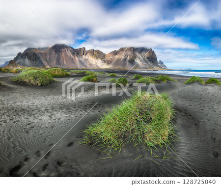 Amazing sunny day and gorgeous rippled black sand beach with dunes and green grass on Stokksnes cape in Iceland. Amazing sunny day and gorgeous rippled black sand beach with dunes and green grass on Stokksnes cape in Iceland. 128725040