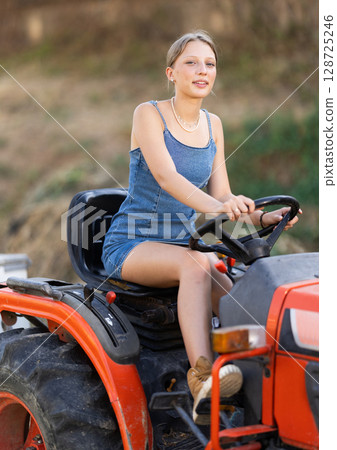 Young woman sits on minitractor and drives rural vehicle, performs loading work on farm Young woman sits on minitractor and drives rural vehicle, performs loading work on farm 128725246