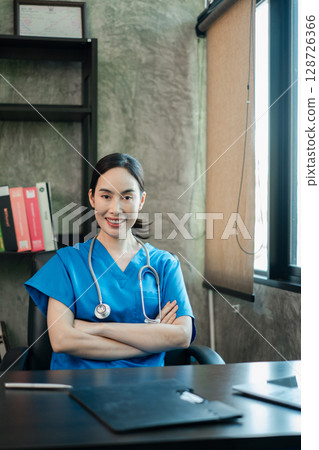 Smiling healthcare worker in blue scrubs with stethoscope, seated at a desk in a contemporary office environment. 128726366