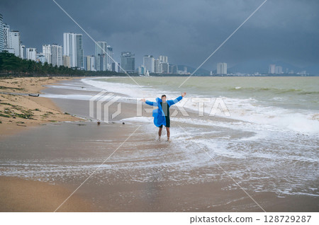 man in a raincoat on the beach by the sea with waves in cloudy weather with rain. Concept of the rainy and monsoon season 128729287