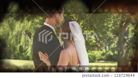 Embracing bride and groom standing in garden wearing wedding dress and tuxedo, with veil and ring Embracing bride and groom standing in garden wearing wedding dress and tuxedo, with veil and ring 128729644