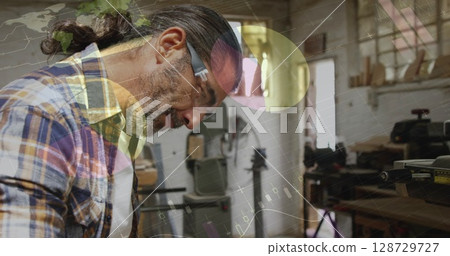 Leaning mature woodworker examining wooden plank on workbench at wood shop, with safety glasses 128729727