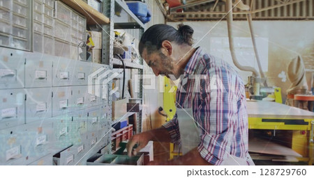 Craftsman wearing apron sorting components from storage drawers in workshop, with yellow machine 128729760