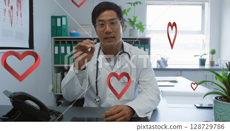 Talking Chinese doctor holding pen and wearing stethoscope in medical room, with smartphone, plants 128729786