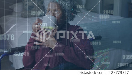Holding ceramic mug senior African American woman in wheelchair gazing in lit bedroom, data overlay 128729817