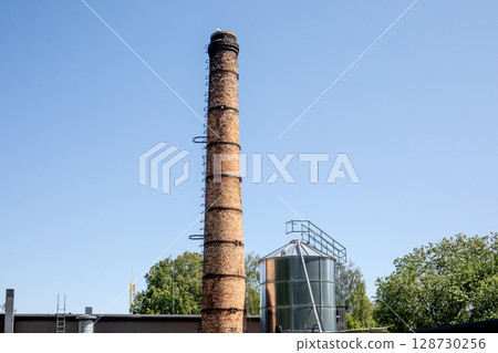 Old Industrial Chimney and Metal Silo Under Clear Blue Sky in Summer Old Industrial Chimney and Metal Silo Under Clear Blue Sky in Summer 128730256