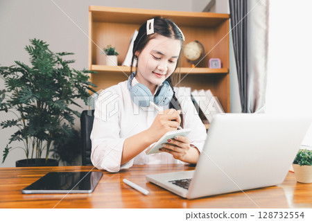 Asian girl student wear headphones learning online watching webinar class looking at laptop computer eLearning remote lesson making notes or video calling virtual conference meeting teacher at home. Asian girl student wear headphones learning online watching webinar class looking at laptop computer eLearning remote lesson making notes or video calling virtual conference meeting teacher at home. 128732554