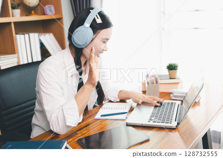 Asian girl student wear headphones learning online watching webinar class looking at laptop computer eLearning remote lesson making notes or video calling virtual conference meeting teacher at home. 128732555