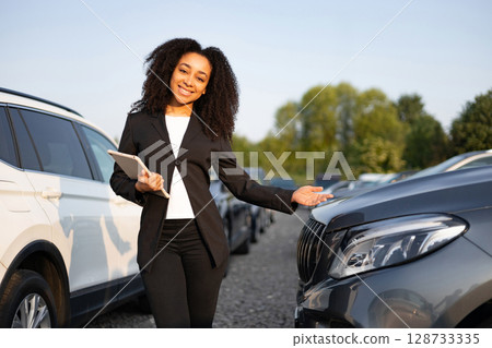 Smiling car salesperson presenting a vehicle on a sunny day. The woman holds a tablet and gestures towards the car. 128733335