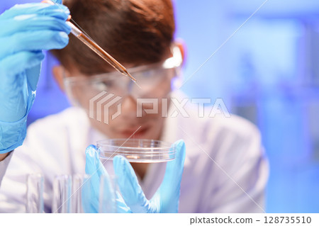 Female lab technician holding pipette above Petri dish. Young scientist doing analysis in the laboratory. 128735510
