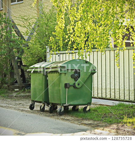green trash containers in sunlit street near fence with birch tree branches and greenery green trash containers in sunlit street near fence with birch tree branches and greenery 128735728