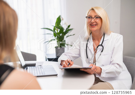Senior family doctor visits patient at home, takes medical history, explains treatment plan. Elderly therapist visiting young woman at home, asking questions, writing notes, discussing therapy options 128735965