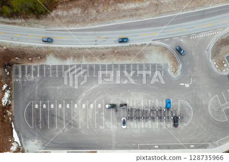 Electric vehicle approaches charging station within parking lot. Grey asphalt with numerous parking lines suggests quiet moment at modern facility 128735966