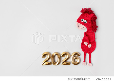 Crocheted toy fiery red horse and gold numbers. The symbol of the year 2026 on a gray background. Crocheted toy fiery red horse and gold numbers. The symbol of the year 2026 on a gray background. 128736654