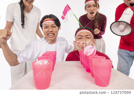 Portrait of an Indonesian southeast asian people celebrate Indonesia Independence Day 17th of August with game blowing ball on plastic glass isolated white background. Concept of Kemerdekaan Indonesia Portrait of an Indonesian southeast asian people celebrate Indonesia Independence Day 17th of August with game blowing ball on plastic glass isolated white background. Concept of Kemerdekaan Indonesia 128737099