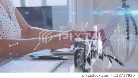 Typing woman wearing white polka-dot blouse on silver laptop at study desk, with tripod camera 128737920