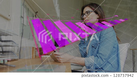 Analyzing woman wearing denim shirt holding tablet at office desk, with monitor and paper trays Analyzing woman wearing denim shirt holding tablet at office desk, with monitor and paper trays 128737942