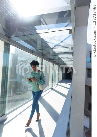 African American woman walking in glass corridor, checking smartphone and carrying tote, copy space 128738469