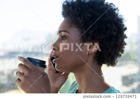African American woman talking on smartphone holding coffee cup in office near window, copy space 128738471