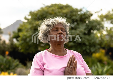Senior woman meditating in garden park with hands clasped at chest, expressing calm focus Senior woman meditating in garden park with hands clasped at chest, expressing calm focus 128738472
