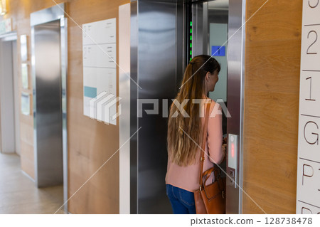 Mid-adult woman standing at open elevator doors in wood-paneled lobby, carrying handbag, copy space Mid-adult woman standing at open elevator doors in wood-paneled lobby, carrying handbag, copy space 128738478
